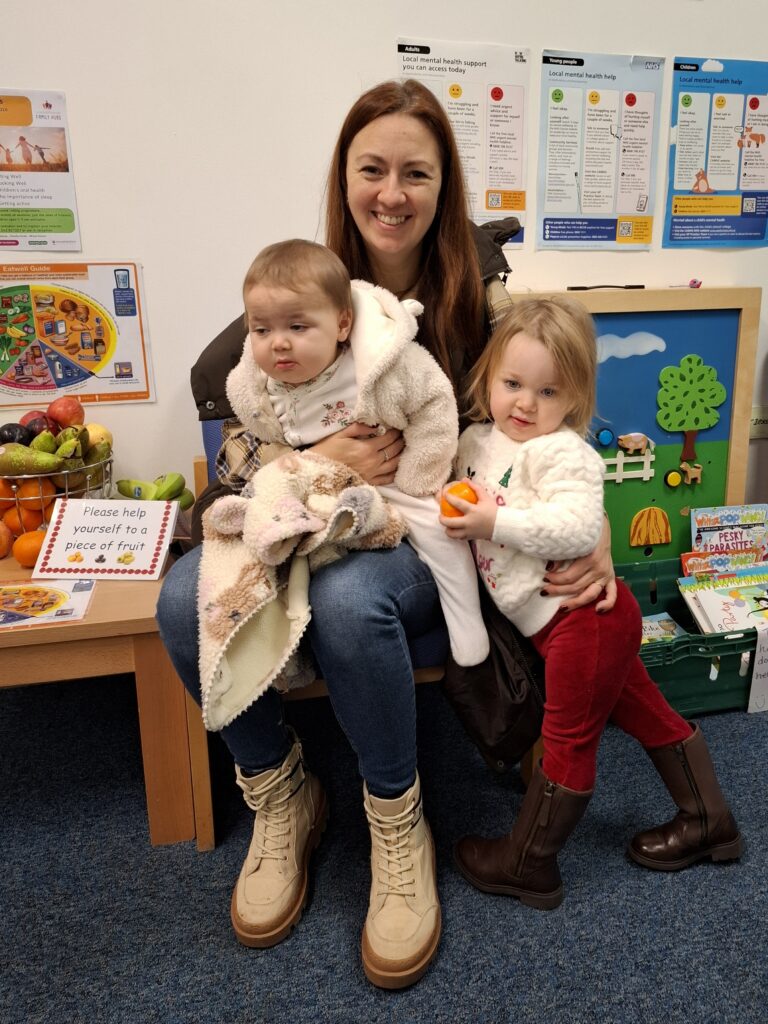 A lady sat down smiling with a baby on her knee and a toddler by her side holding an orange. To the left is a bowl of fresh fruit and healthy eating info.