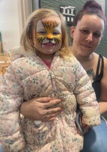 A young child with a painted tiger face smiles at the camera while being held by an adult indoors. The face paint features orange, white and black stripes with whiskers and small fangs. The child wears a light coloured coat with a floral pattern. The adult stands close behind, holding the child securely. In the background, there is a wall with a light switch panel and part of a room with furniture.