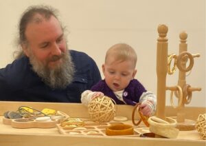 A baby sits at a low wooden play table with natural toys including rings, a wicker ball and small objects. The baby reaches forward to explore the items. An adult sits close behind, watching and supporting. The play area features wooden activity frames with hanging rings on the right. The setting appears to be an indoor early years space with neutral walls and simple furniture focused on sensory play and exploration.